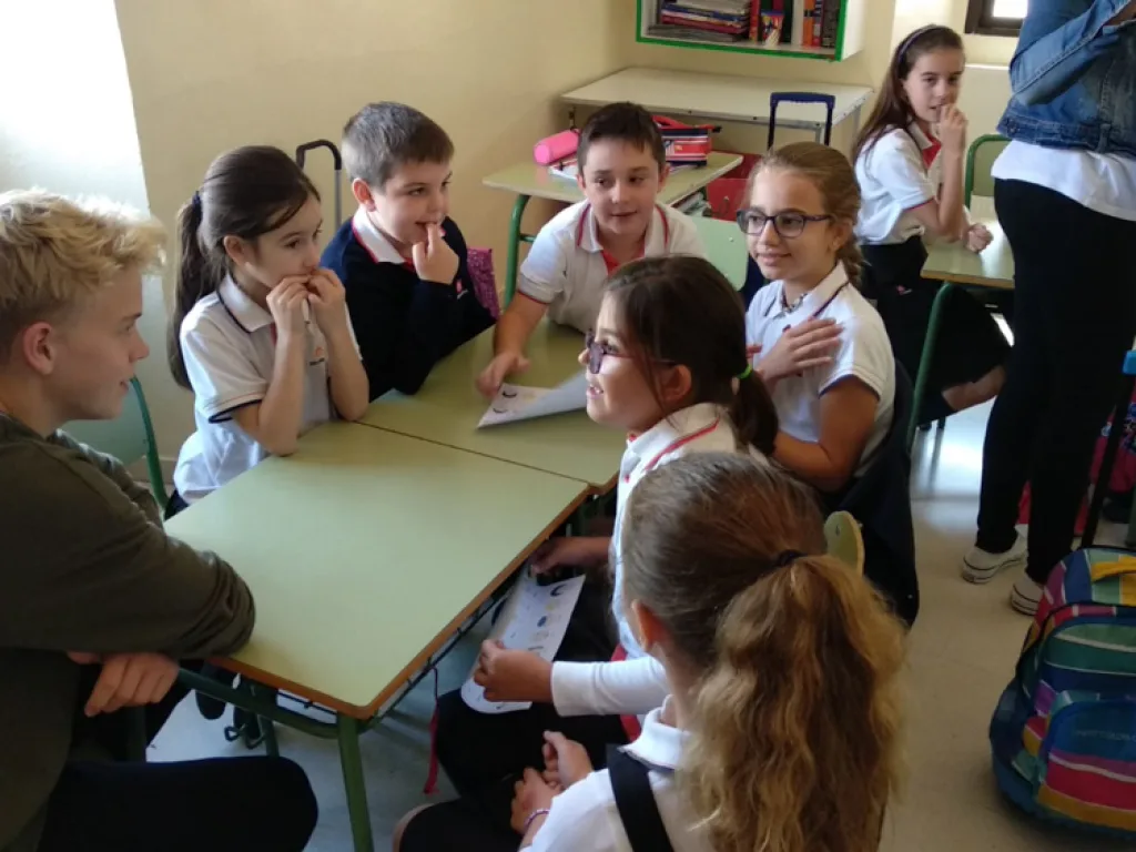 Jugendliche sitzen mit spanischen Schulkindern an Tischen im Klassenraum.